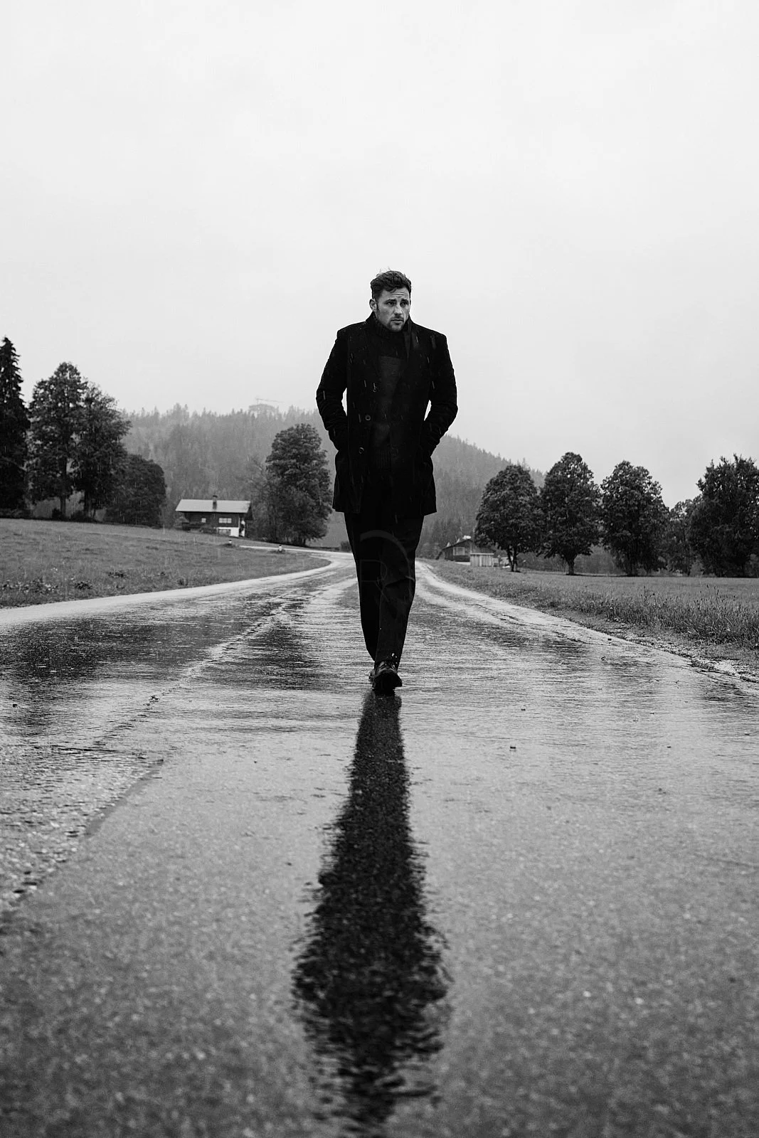 A man walking alone on a wet, empty road in a rural area with trees and houses in the background, black and white photograph.