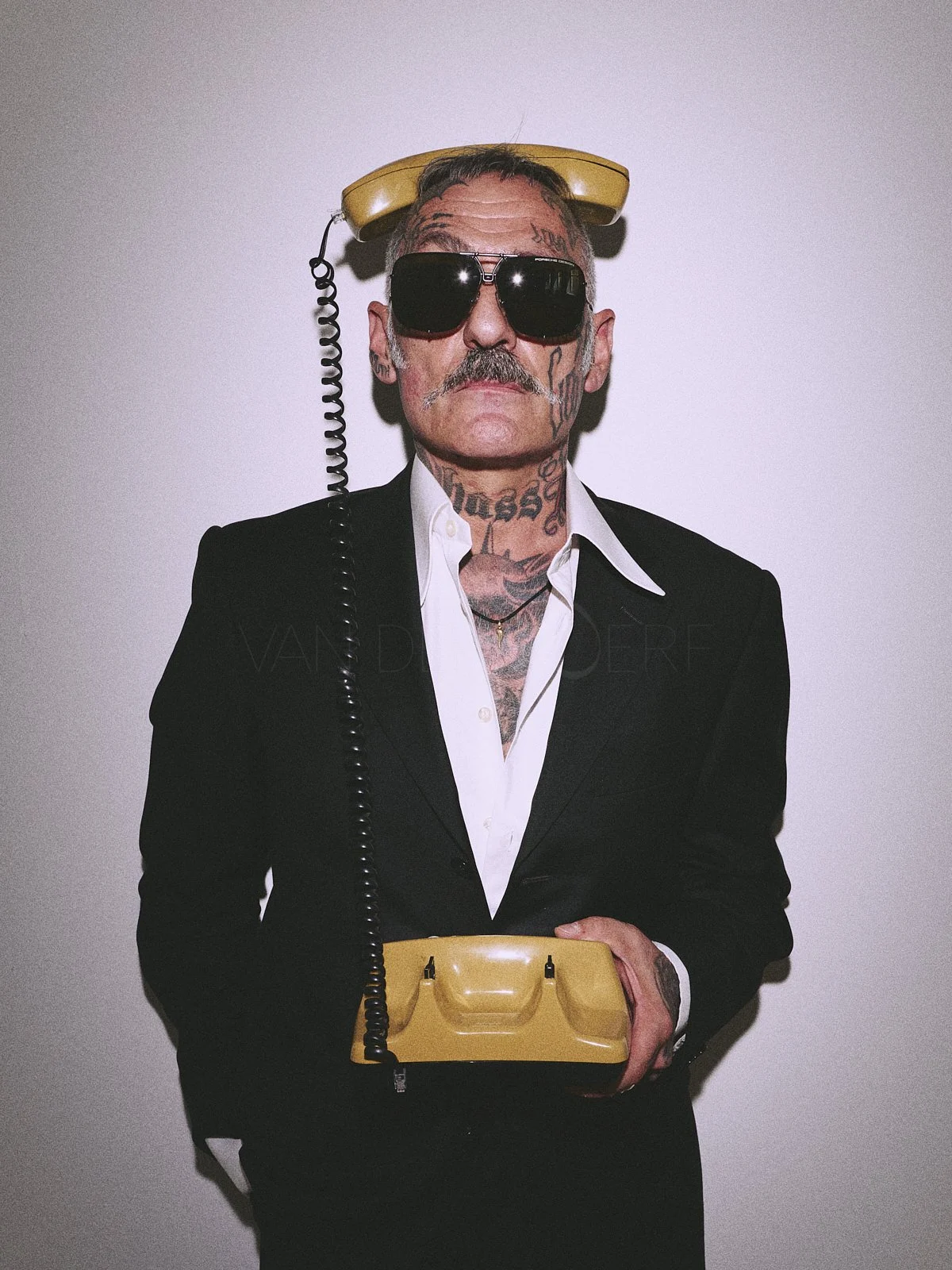 Man with tattoos, wearing black suit and white shirt, dark sunglasses, holding a yellow vintage telephone with a telephone receiver on his head, against a plain white wall.