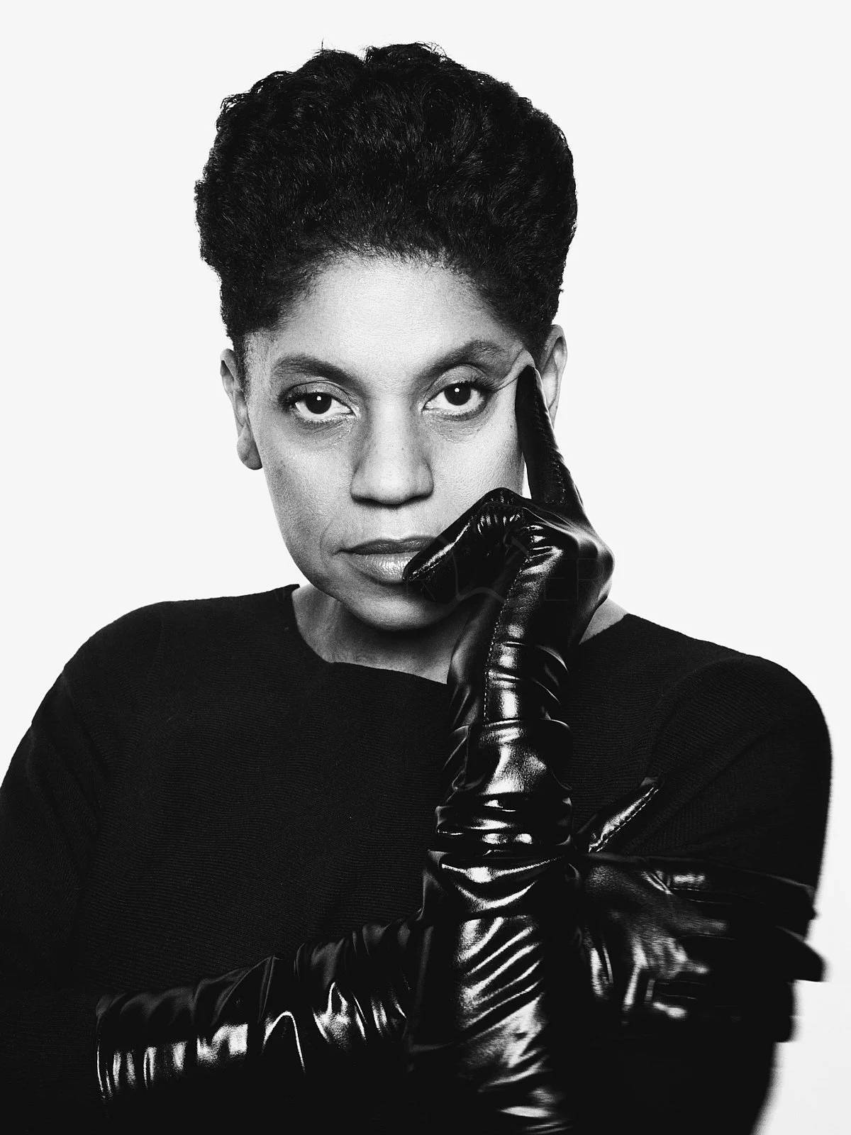 Black and white portrait of a woman with short, textured hair, wearing a black top and long, shiny black gloves, with one finger resting on her temple, looking directly at the camera.