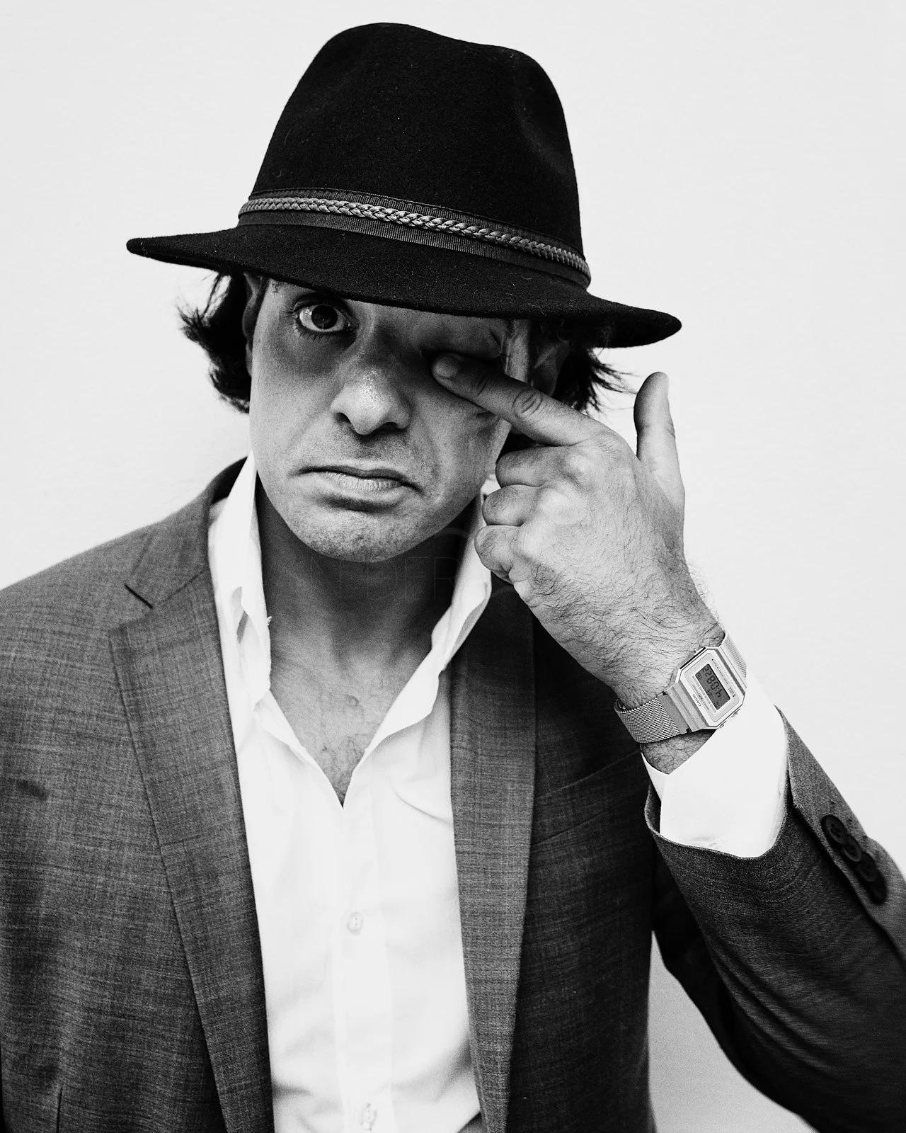 A man in a suit wearing a hat, pulling his eye with his fingers, looking directly at the camera.