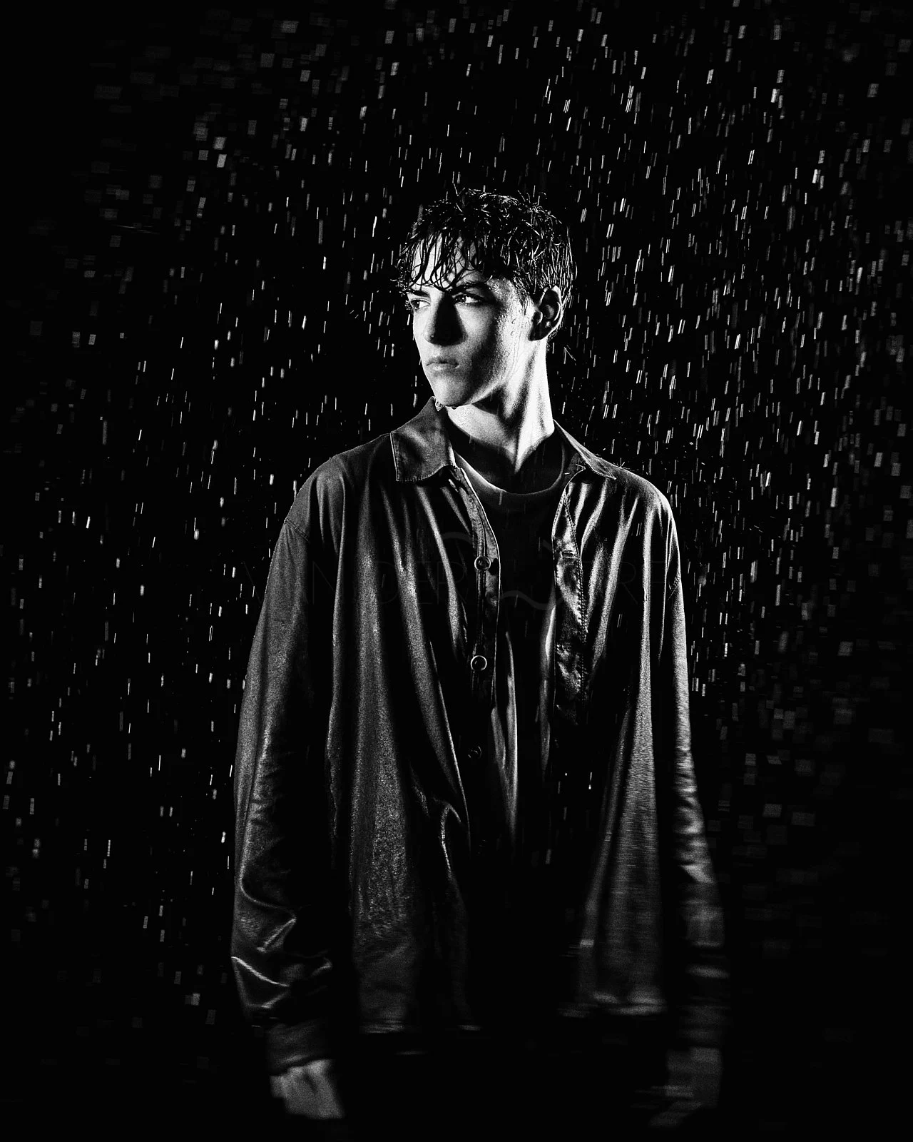 Black and white photo of a young man in a raincoat standing in the rain, looking to his left, with water droplets falling around him.
