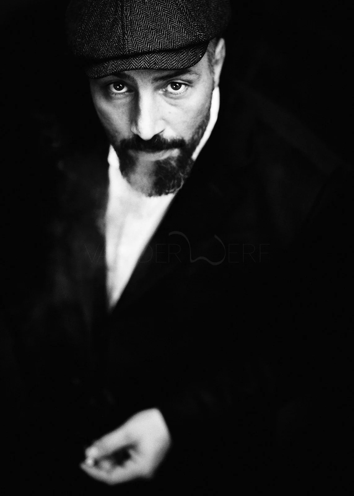 Black and white portrait of a man wearing a tweed flat cap, looking directly at the camera with a serious expression, in low lighting.