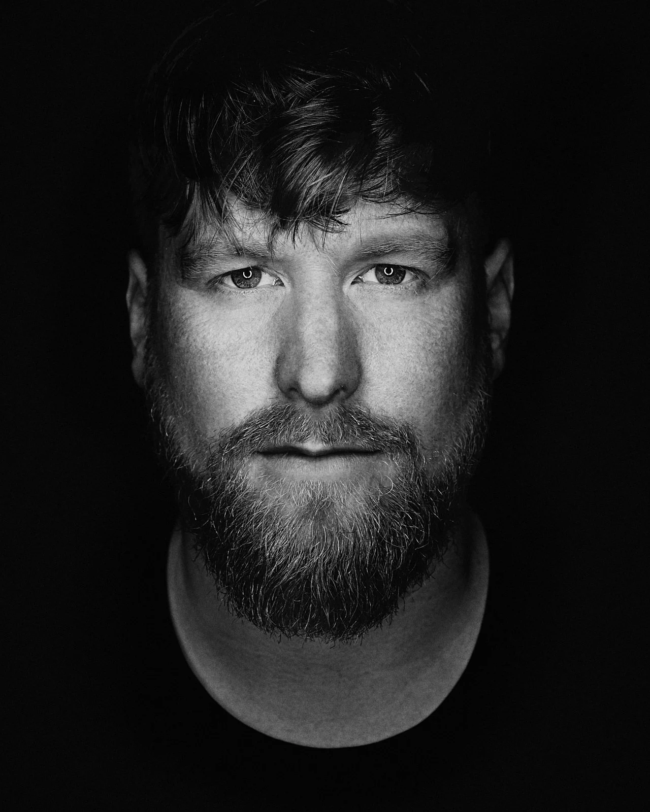 Black and white close-up portrait of a man with a full beard, mustache, and medium-length hair, looking directly at the camera with a neutral expression.