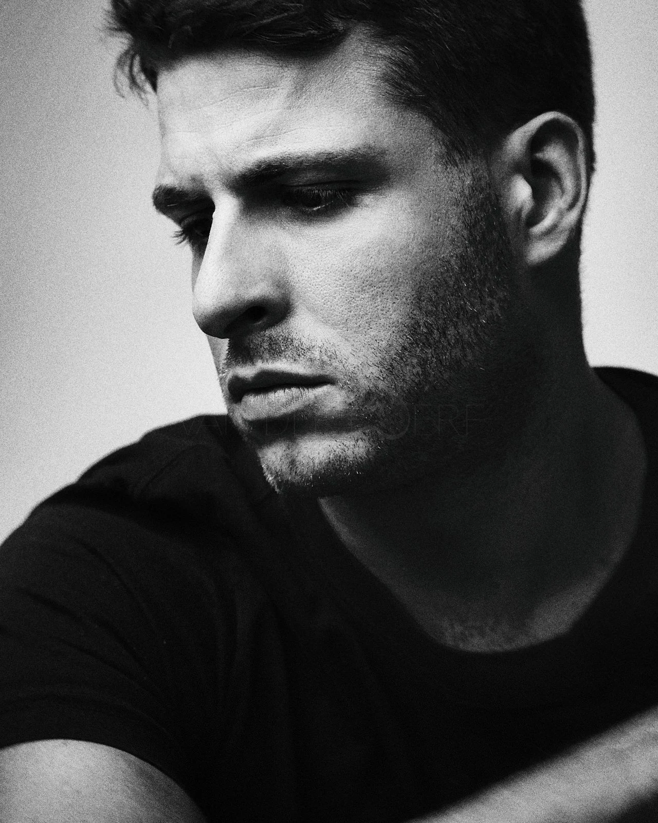 Black and white close-up portrait of a young man with a serious expression, looking downward and to the side, wearing a dark shirt.