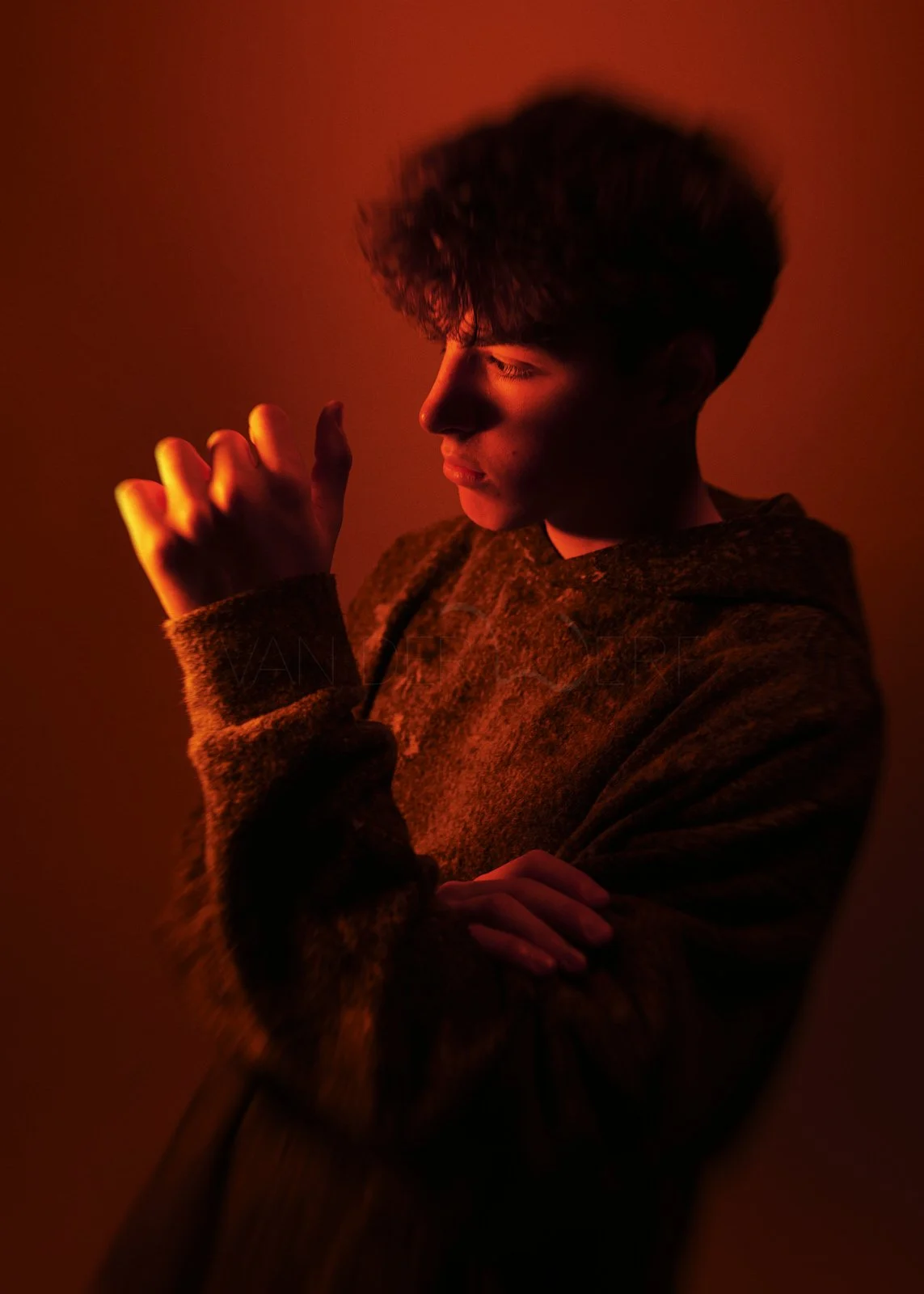A young man with dark hair in a hoodie, illuminated by red light, looking down with his eyes closed and hands folded together.