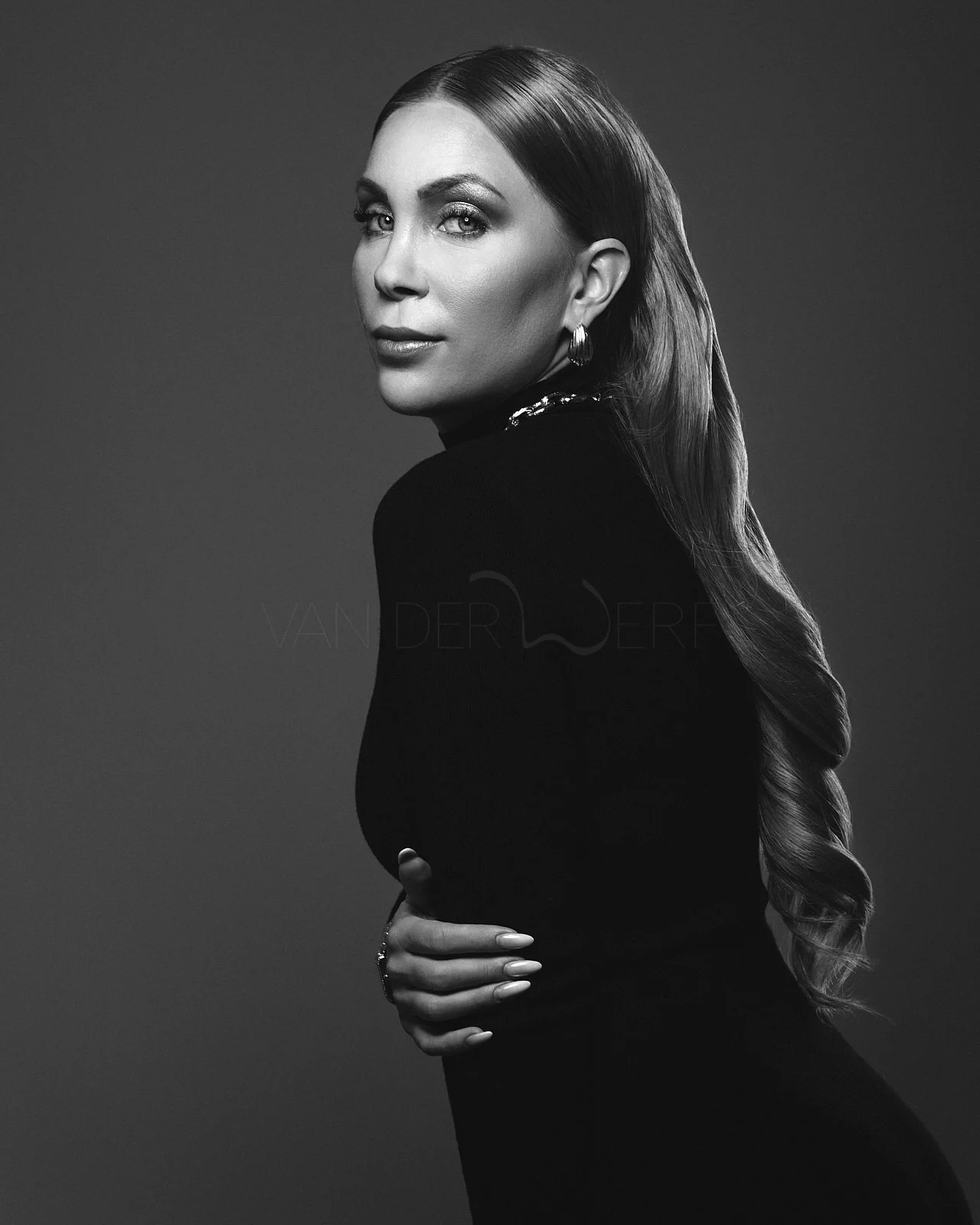 Black and white portrait of a woman with long wavy hair, wearing earrings, and dressed in a long-sleeved black top, posing with one hand on her waist against a gradient background.