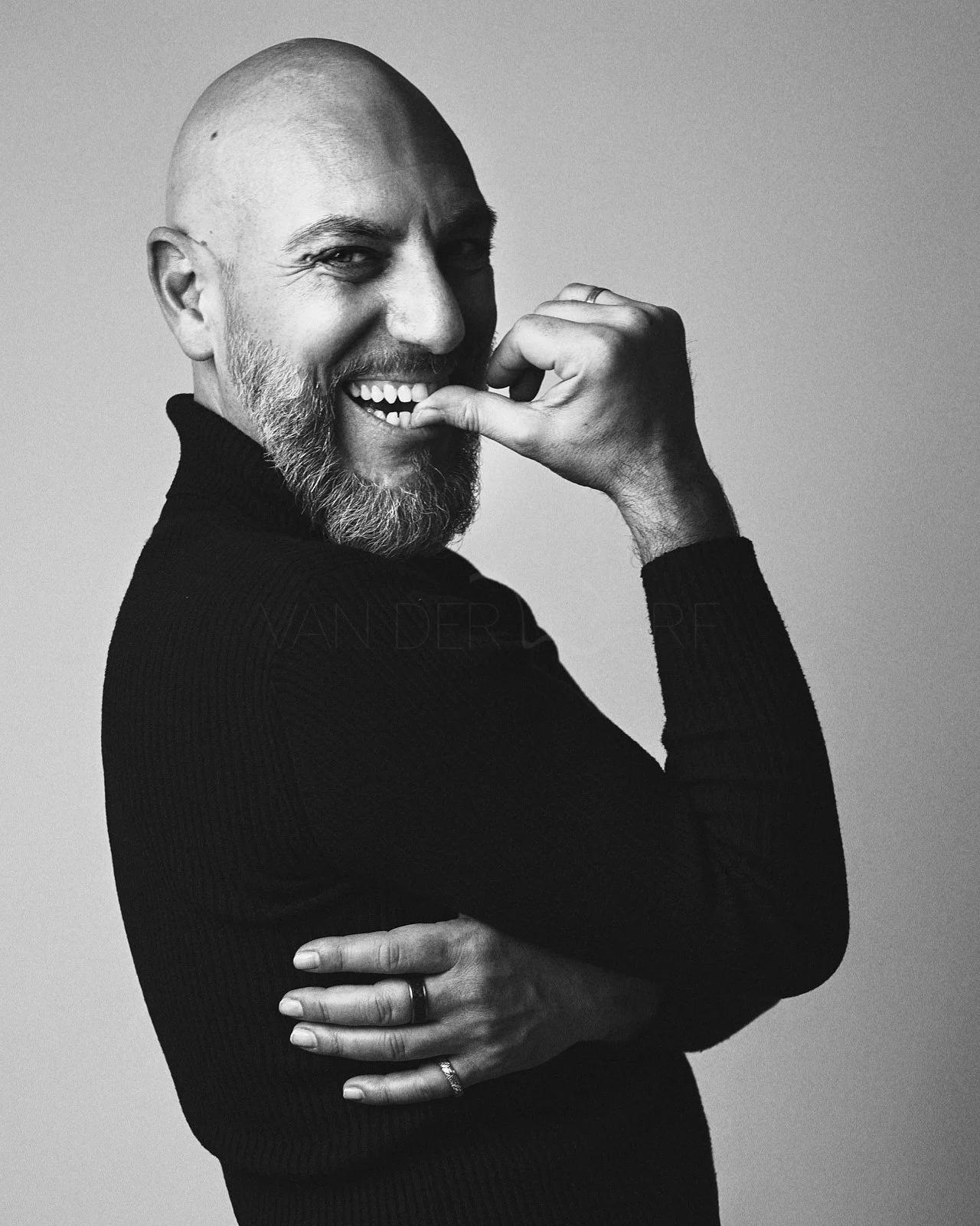 Black and white portrait of a smiling man with a beard, wearing a dark sweater, holding a finger to his mouth, with his other arm crossed over his chest, against a plain background.