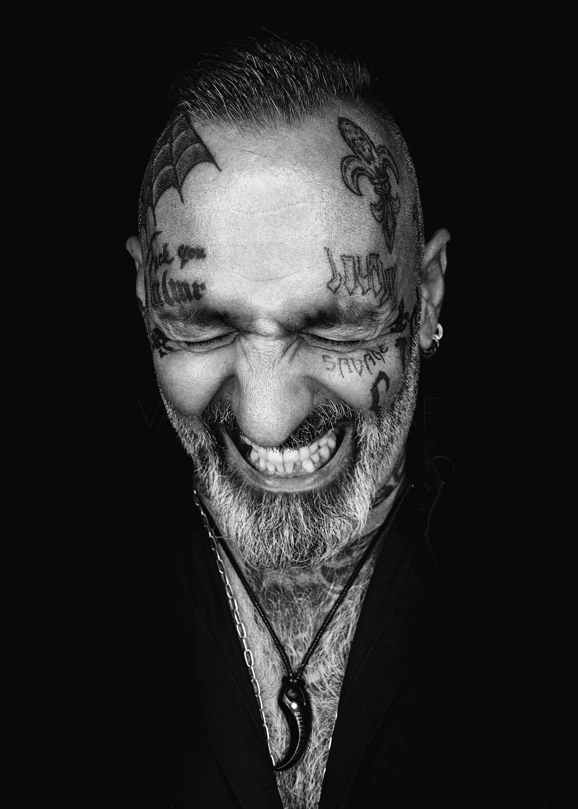 A black and white portrait of a man with tattoos on his face, laughing with his eyes closed, showing his teeth, wearing necklaces and earrings, with a dark background.
