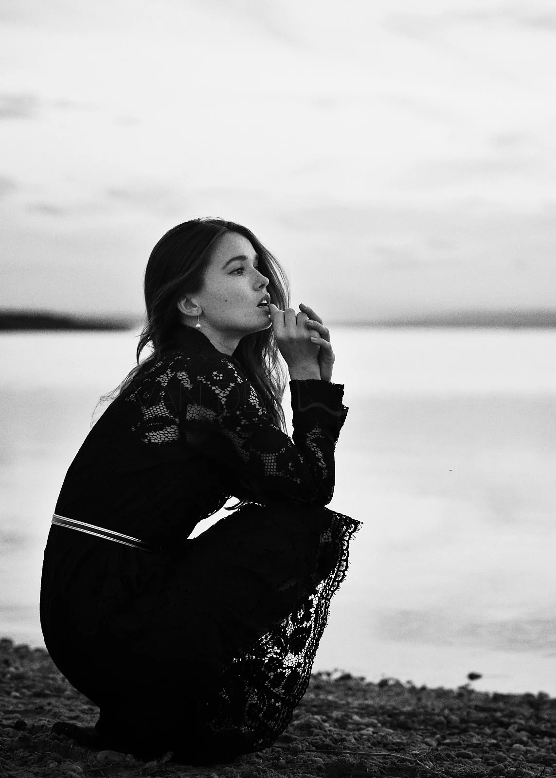 A woman, dressed in black lace clothing, is crouching on a beach near a body of water. She has long hair and is looking to the side with her hands near her face. The image is black and white.