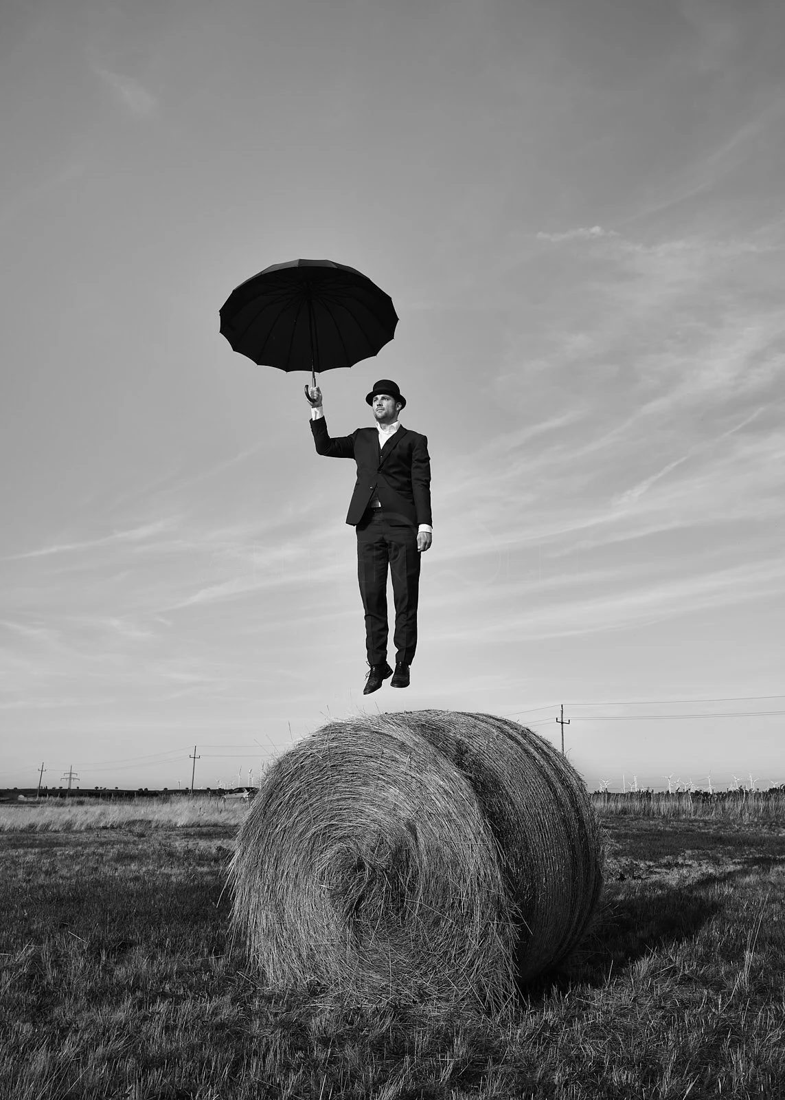 A man in a suit and bowler hat is jumping in the air while holding an umbrella above his head in a field with hay bales. The image is black and white.