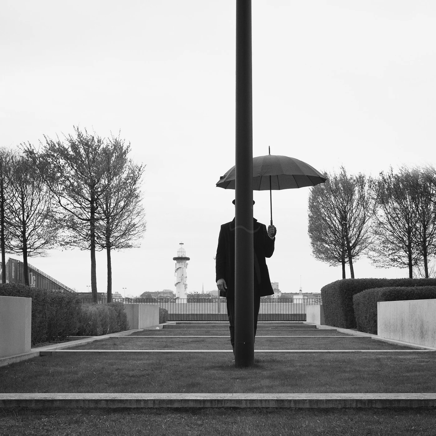 Black and white photo of a person holding an umbrella, standing behind a pole in an outdoor setting with trees and a lighthouse in the background.
