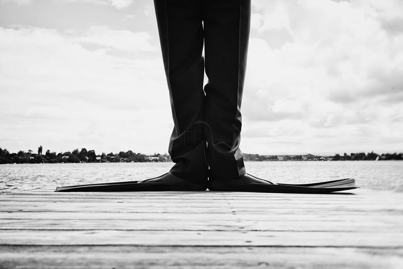 Person wearing business pants and shoes standing on a surfboard on a wooden dock by a body of water, with a cloudy sky in black and white.