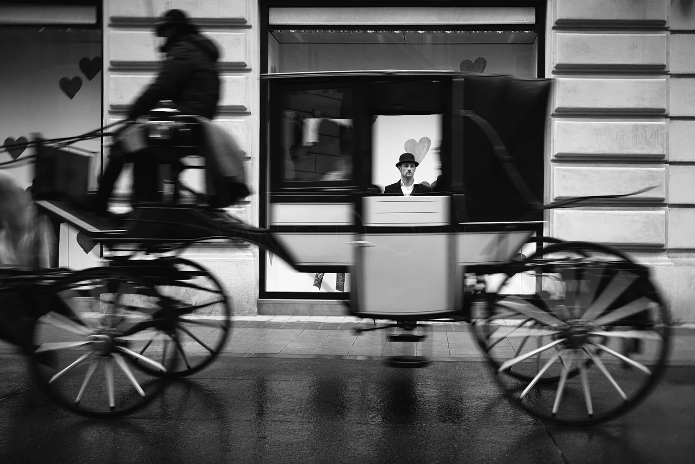 A man wearing a hat and suit looking out of a window behind a horse-drawn carriage on a city street. The photo is in black and white.