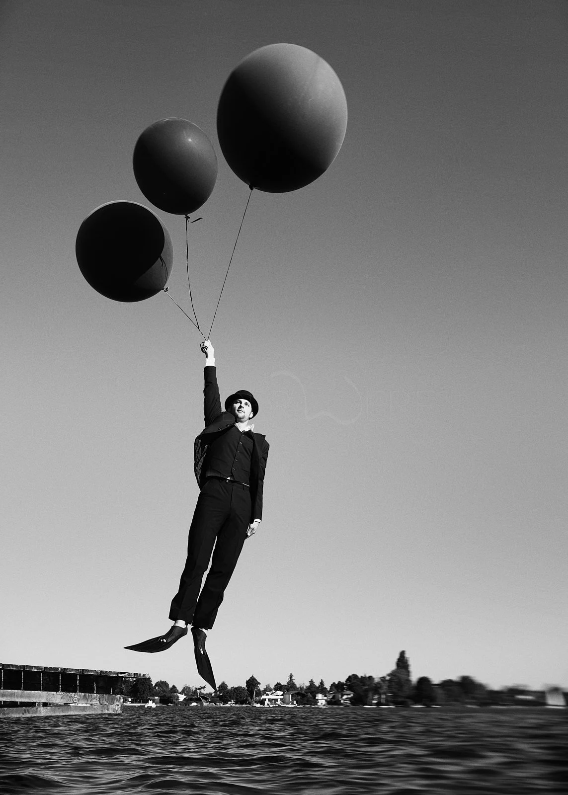 A black and white photo of a person in formal attire, with jet legs, holding three large balloons and floating above water with a cityscape in the background.
