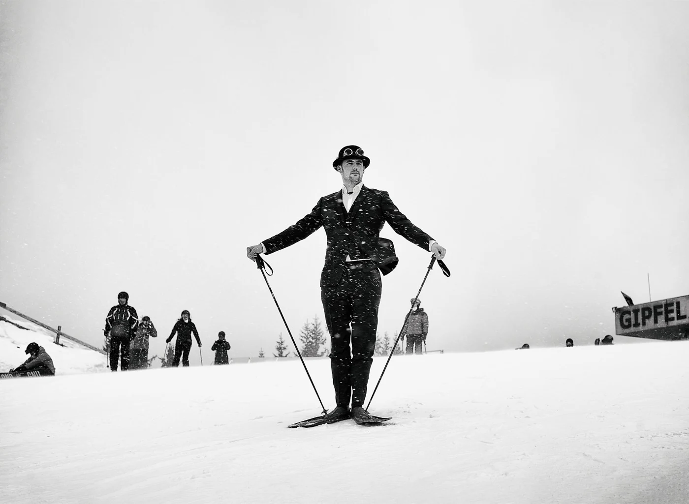 A man in a suit and hat skis on a snowy mountain with a group of people in the background and a sign that reads "GIPFEL".