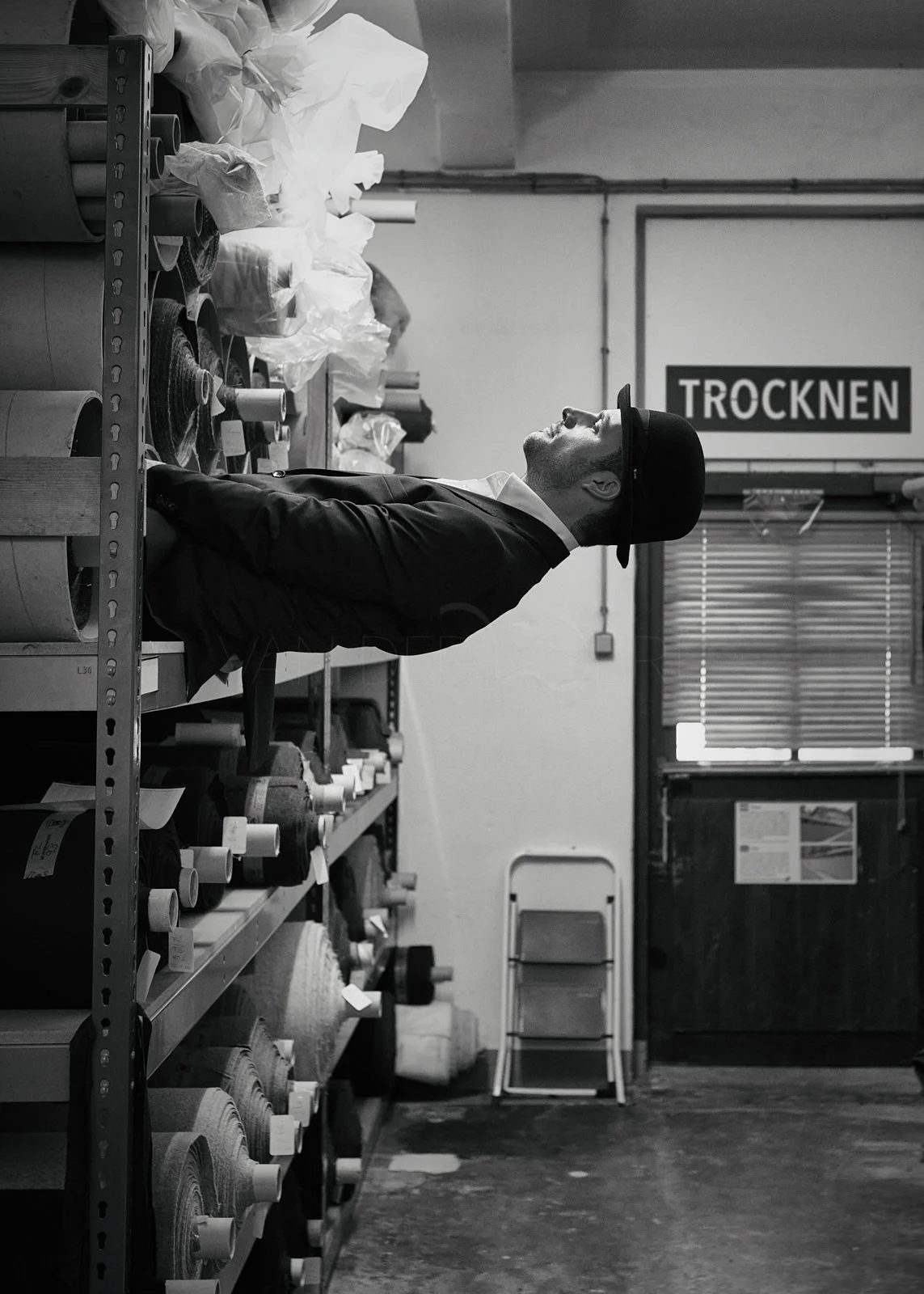 A man in a hat and coat standing in a store aisle filled with rolls of fabric or paper, looking upward. The store has a sign that says 'TROCKNEN' in the background.