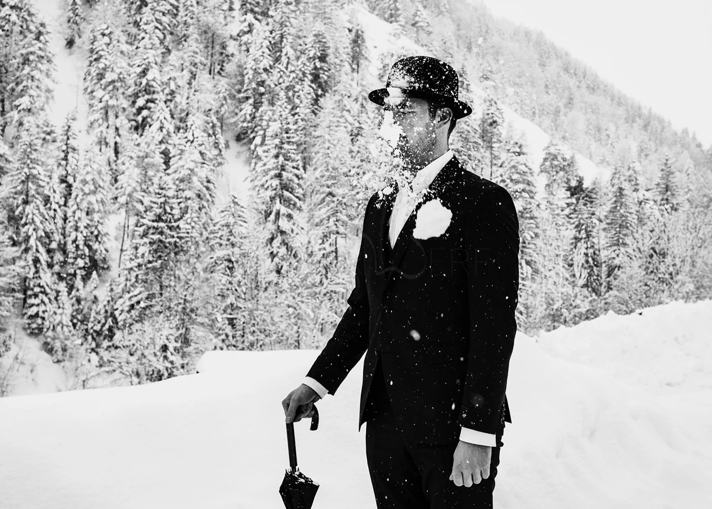 Man in a suit, hat, and sunglasses standing in a snowy forest, with snow on his face and holding a small shovel.