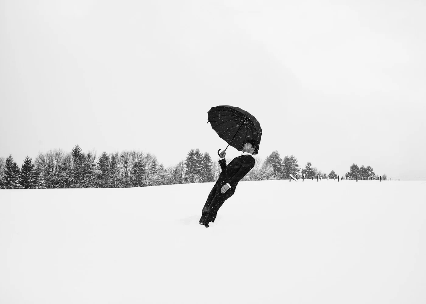 Person in formal attire holding a black umbrella standing in a snowy landscape with a line of trees in the background.