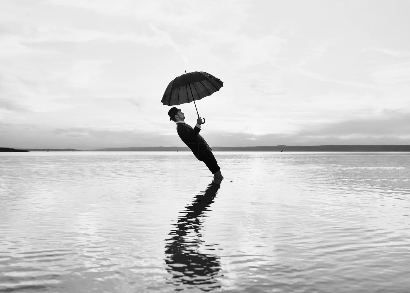 A person dressed in a suit and hat standing knee-deep in water, holding an umbrella, with their head tilted back and looking upwards, over a calm body of water with a distant horizon and cloudy sky.