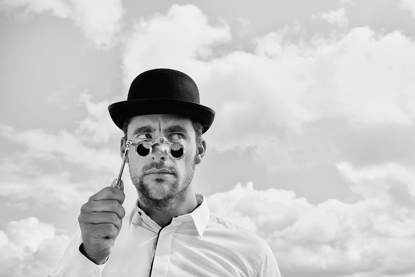 Man wearing a bowler hat and white shirt, holding a lorgnette and looking with curiosity at the camera, with a cloudy sky in the background.