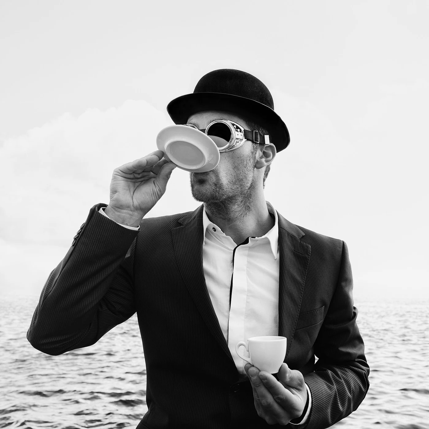 Black and white photo of a man in a suit drinking from a cup with a toy tea set, outdoors by a body of water, wearing a bowler hat and goggles.