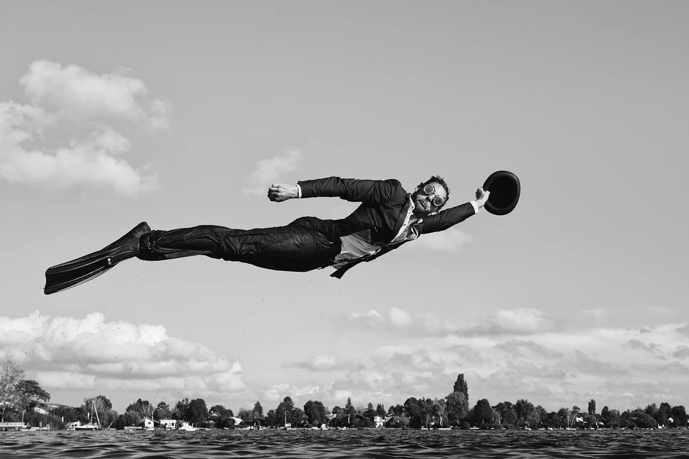 A man in a suit and goggles flies over water holding a hat in the air during daytime, with trees and houses in the background.