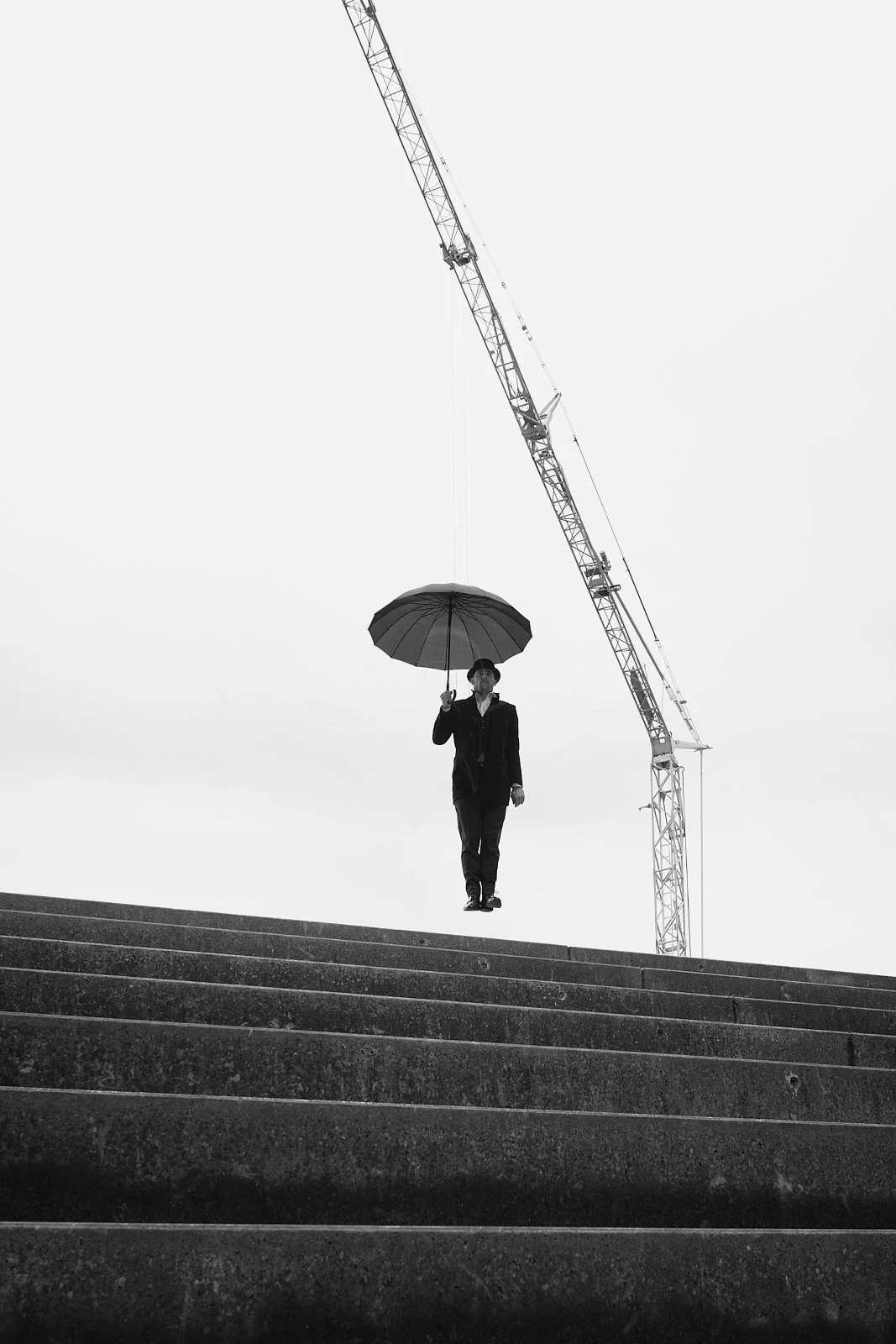 A person in formal attire holding an umbrella while walking down steps, with a large construction crane in the background on a cloudy day.