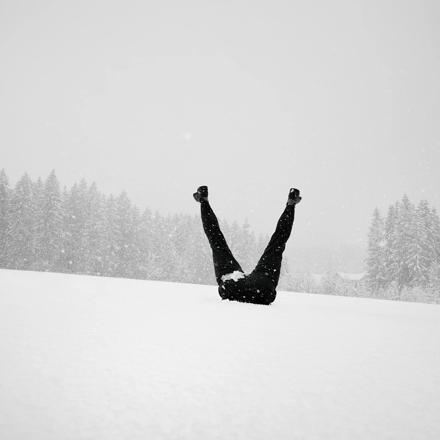 Person lying on the snow with legs raised in the air during snowfall in a snowy landscape.