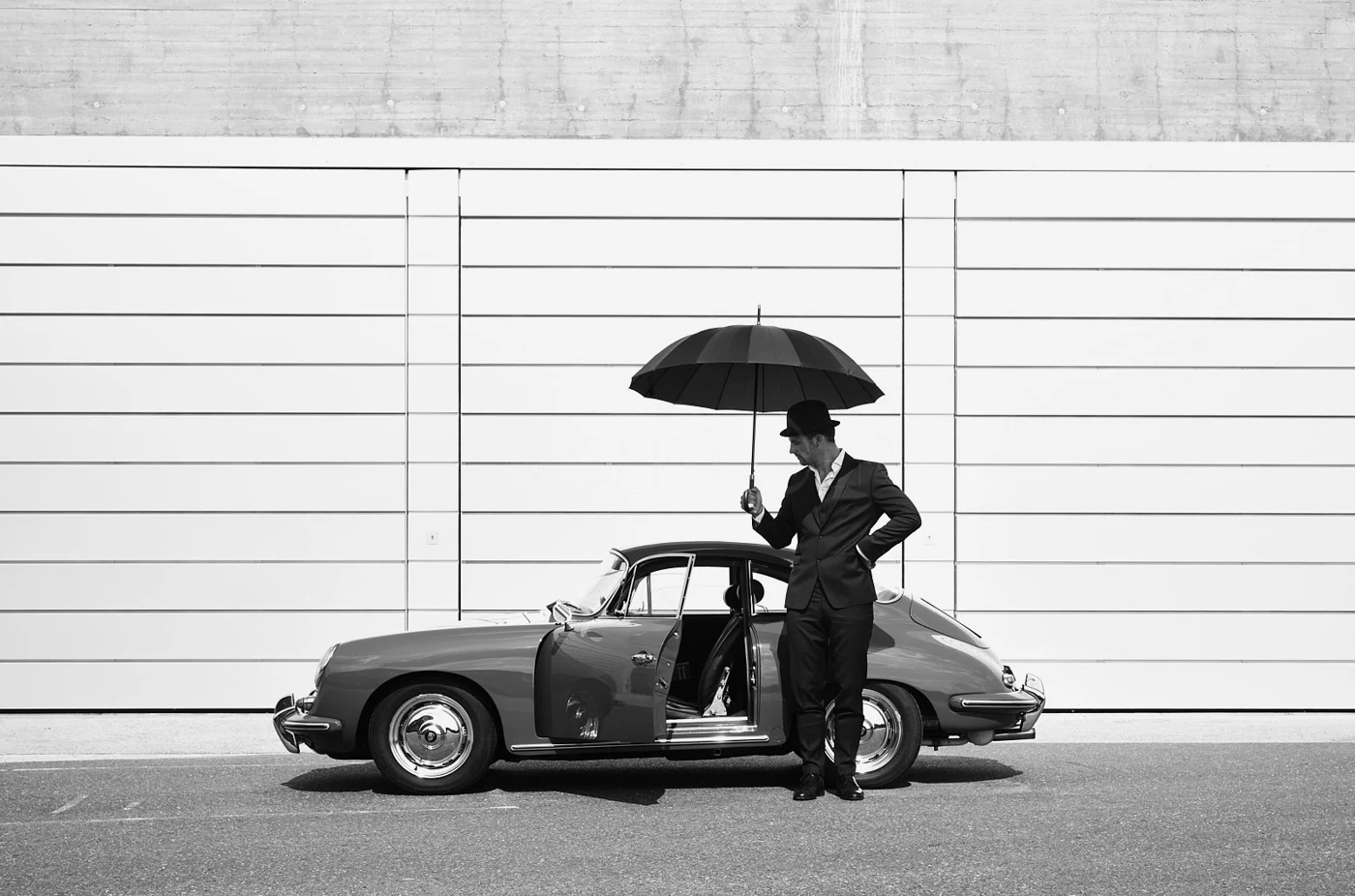 A man in a suit and bowler hat stands beside a classic vintage car, holding an umbrella over his head, against a large garage door with concrete wall above, in black and white.