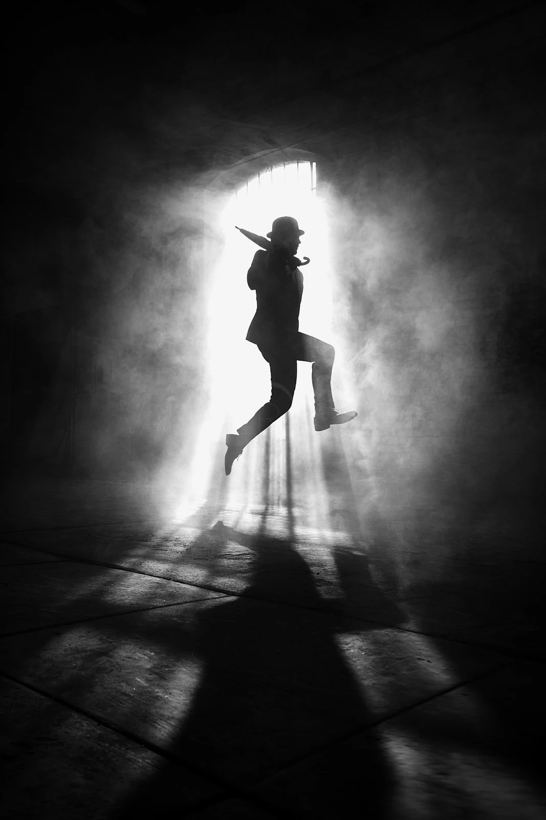 Silhouette of a person jumping in front of a bright light coming through an arched window, surrounded by mist or smoke, creating a dramatic black and white scene.