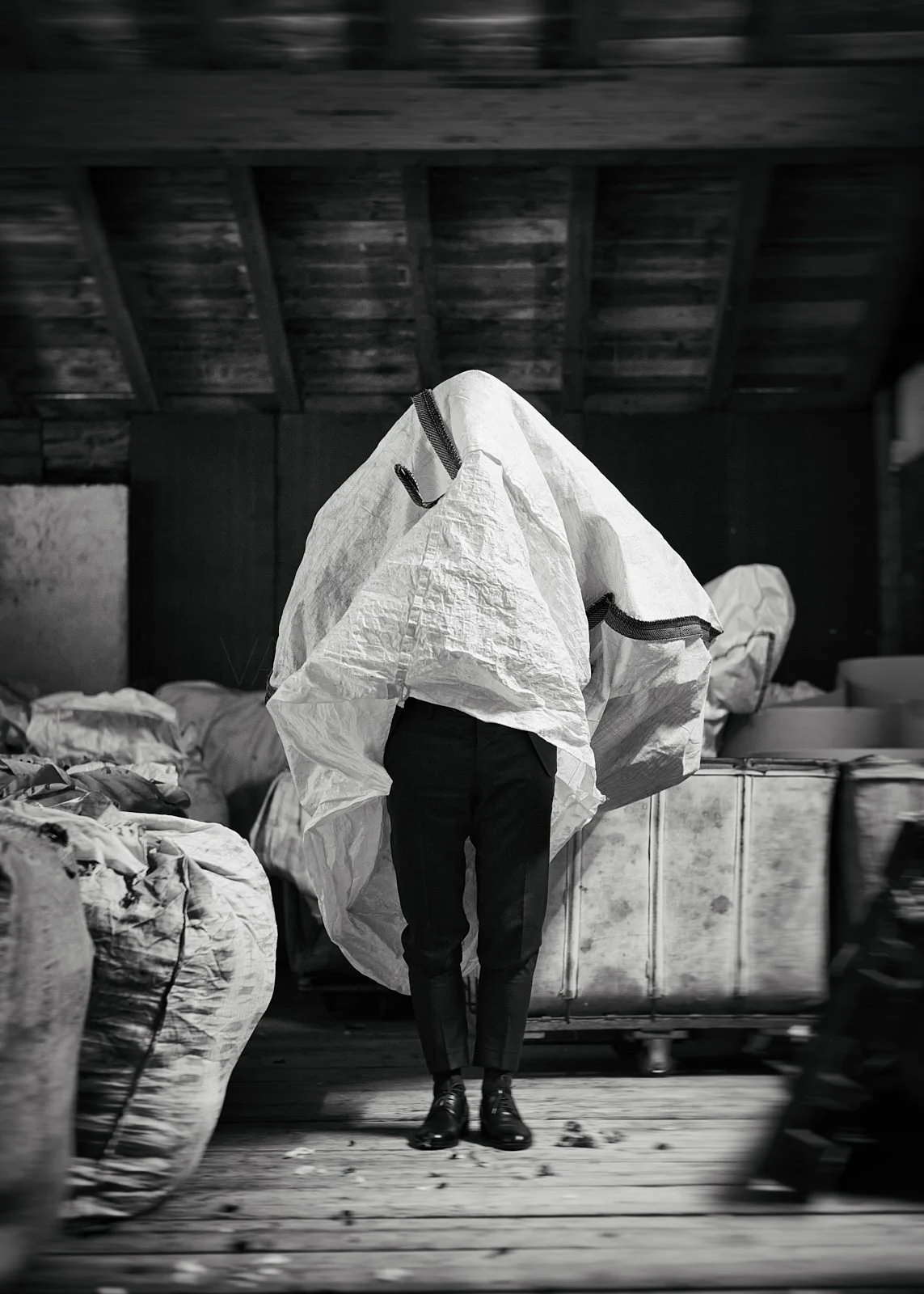 A person dressed in formal shoes and pants is carrying a large object wrapped in a white fabric or material, obscuring their face and upper body, in an industrial or warehouse setting with bags or sacks on the sides.