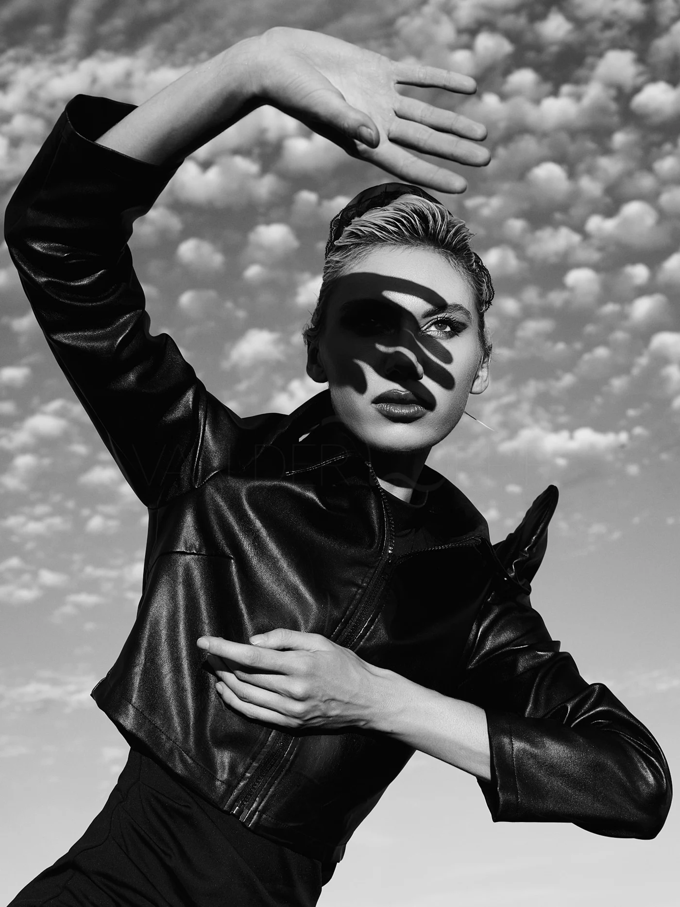 A person in a dark jacket framing their face with raised arms against a cloudy sky.