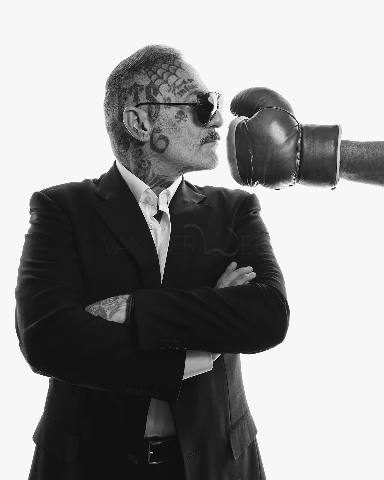 A man in sunglasses and a suit facing a boxing glove with his arms crossed.