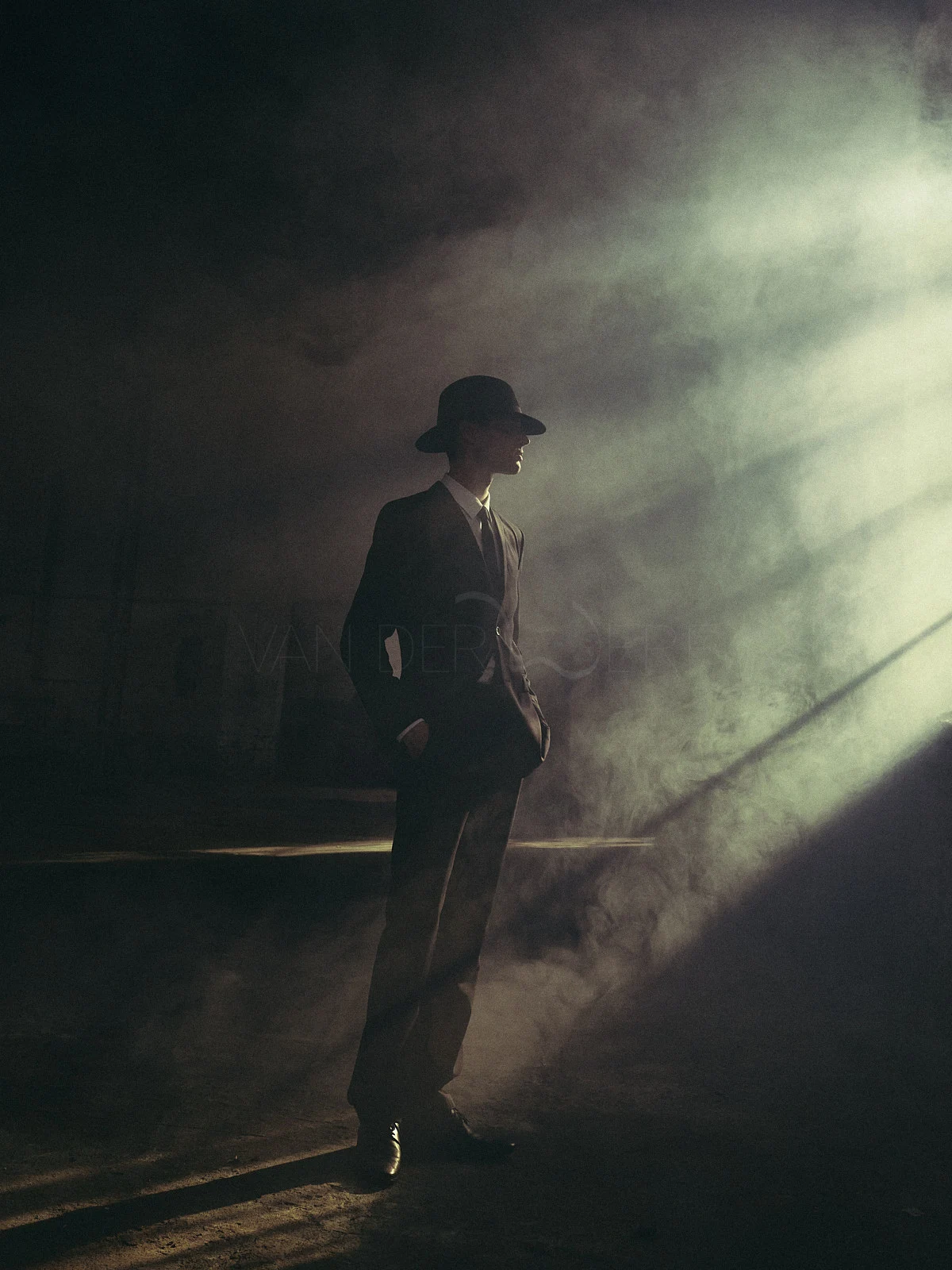 A figure in a hat standing in soft, diagonal rays of studio light.
