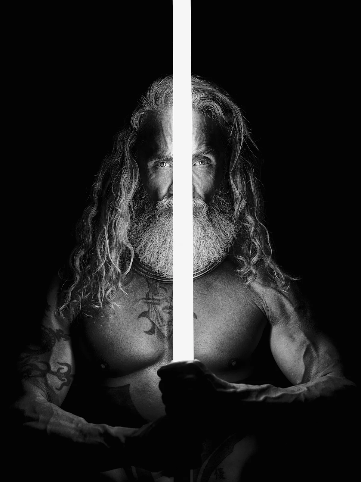 A long-haired man holding a vertical illuminated sword in a dark studio.
