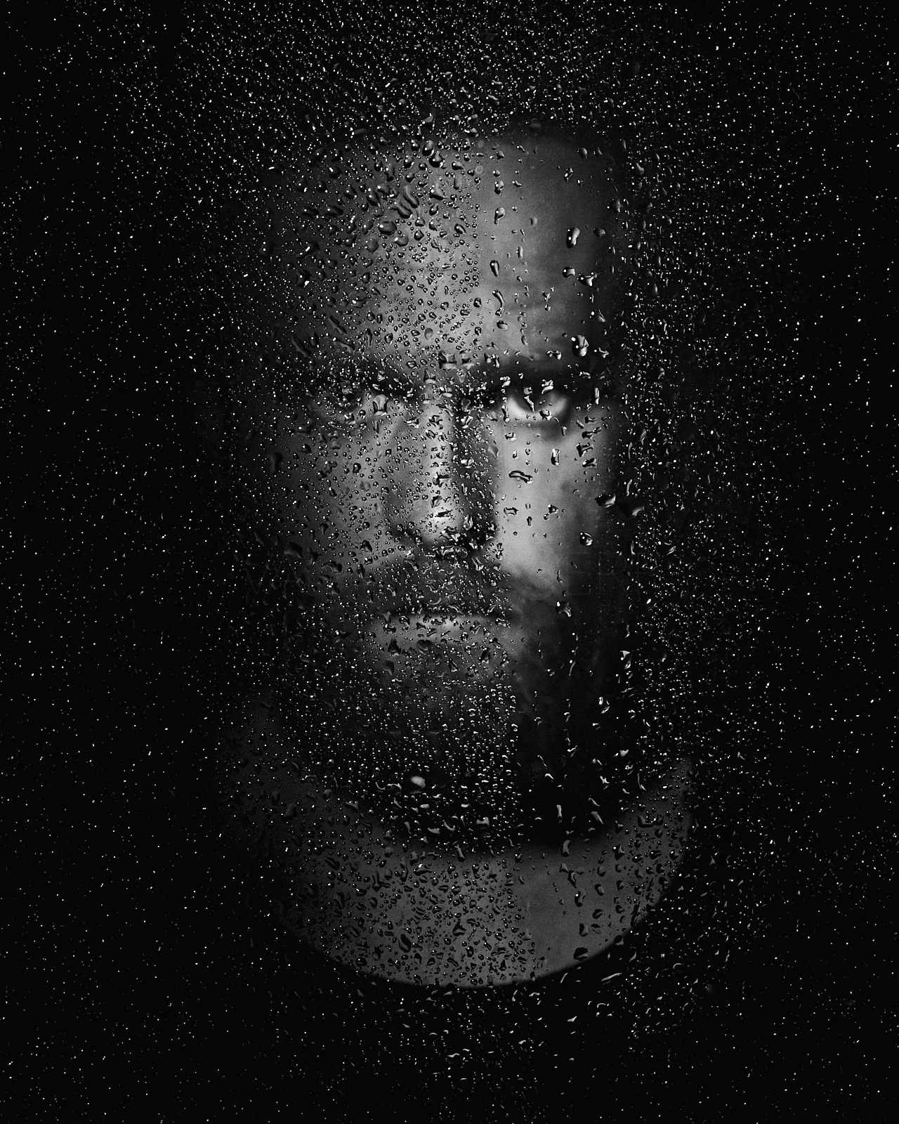A bearded man's face behind a surface covered in droplets.