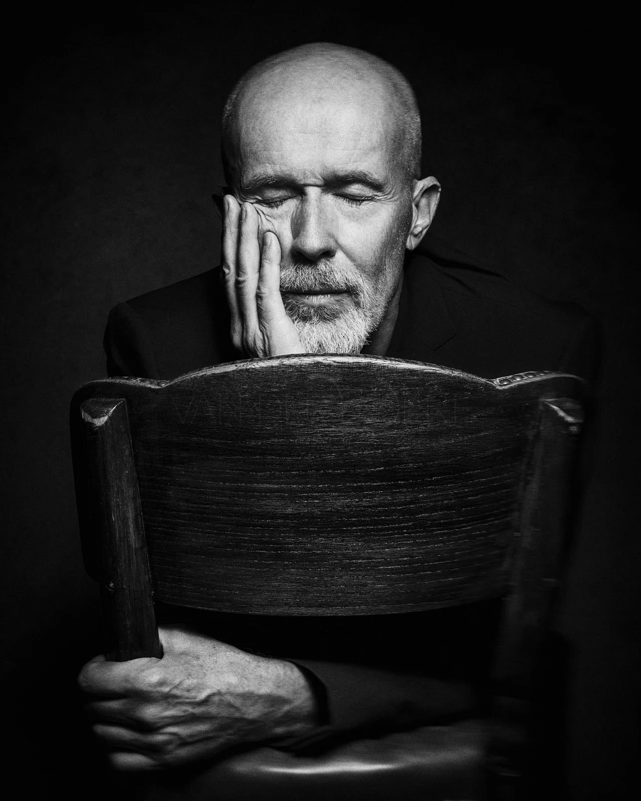 A seated man with closed eyes resting his face in his hand behind a wooden chair.