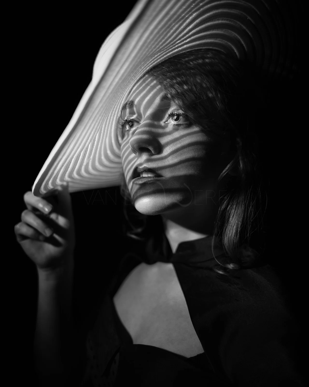 A woman holding a wide-brimmed hat that casts striped shadows across her face.