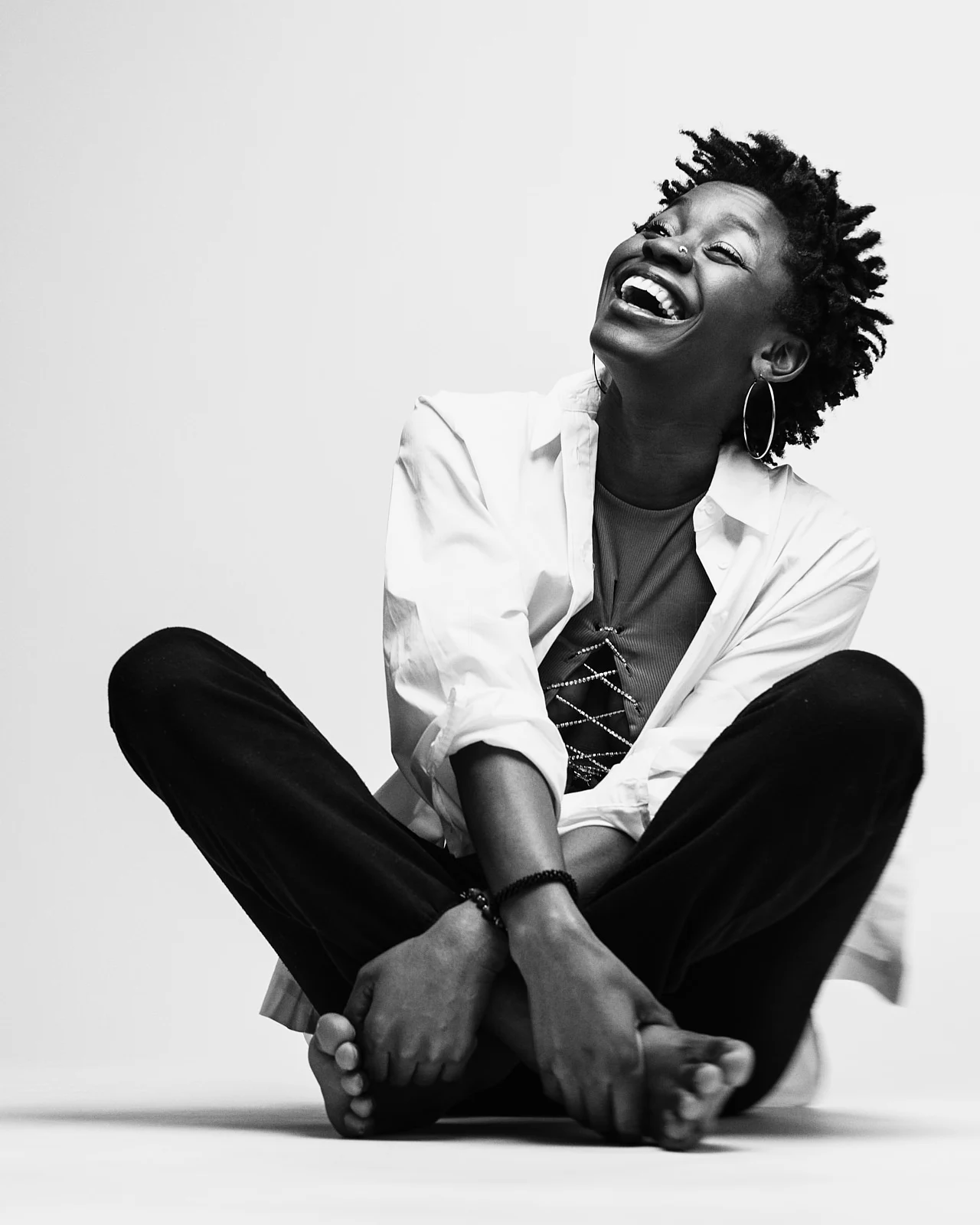 A laughing woman with curls seated cross-legged in a white shirt.