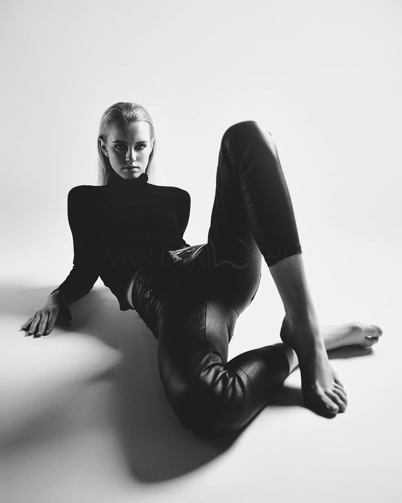 A woman seated on the floor in black clothing and glossy trousers.