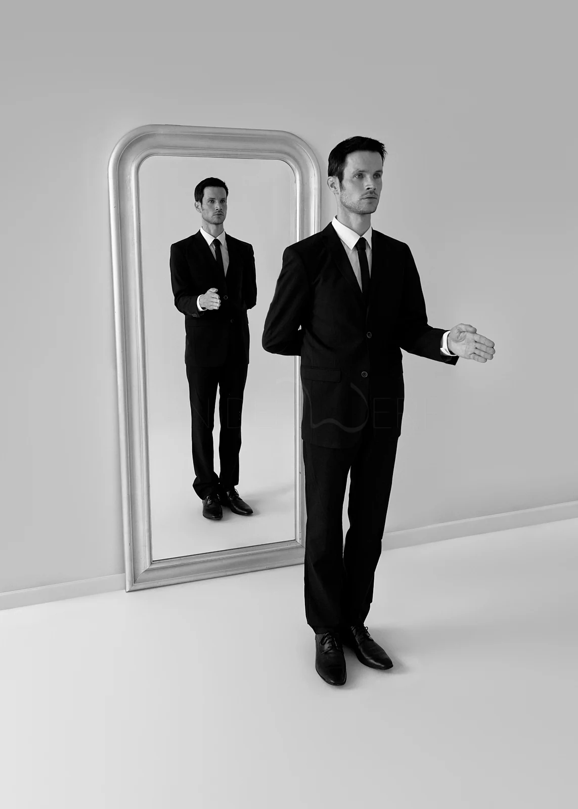 A man in a suit standing beside a tall mirror with his reflection.