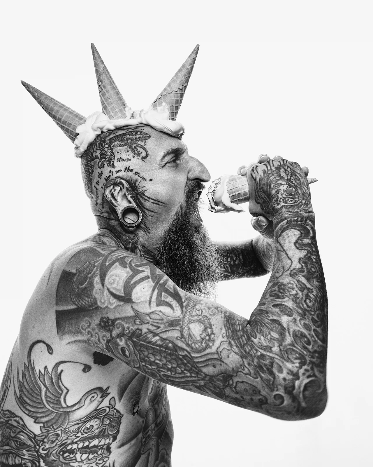 A tattooed man wearing cone-shaped headwear while eating an ice cream.