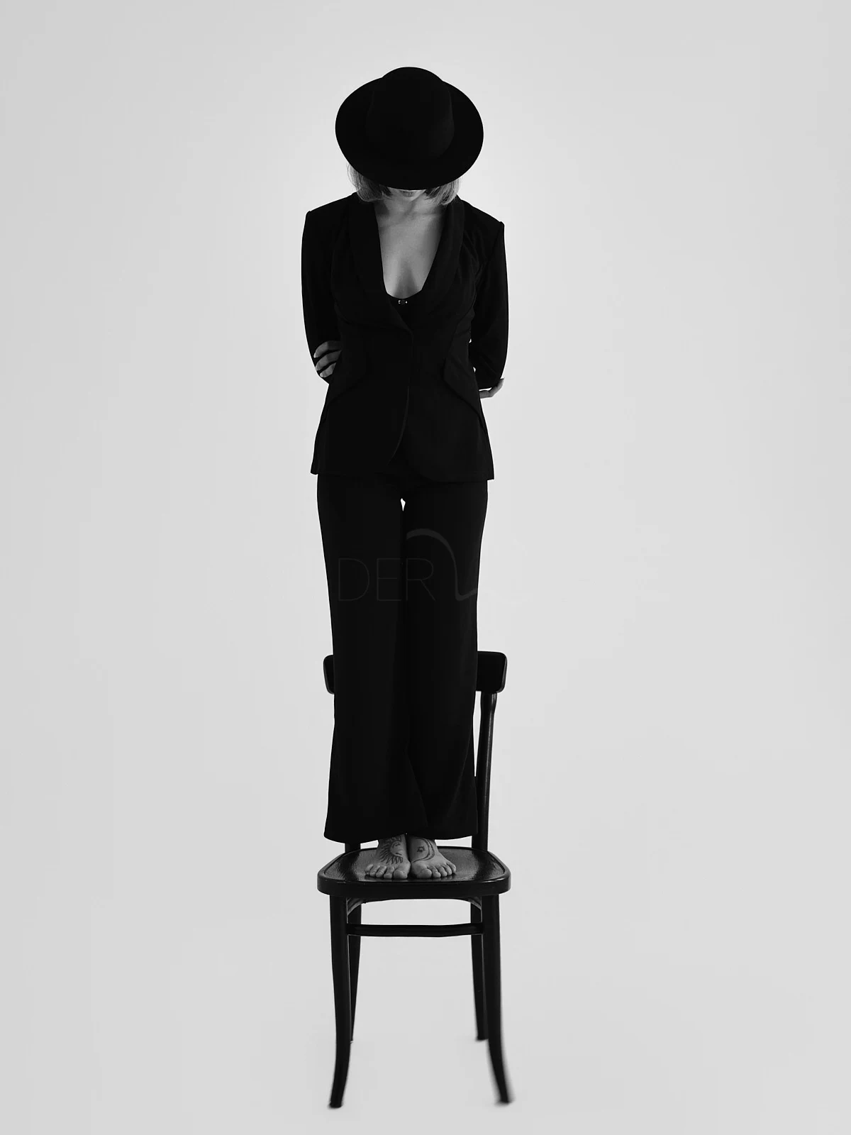 A figure in a dark suit and hat standing on a chair with head lowered.