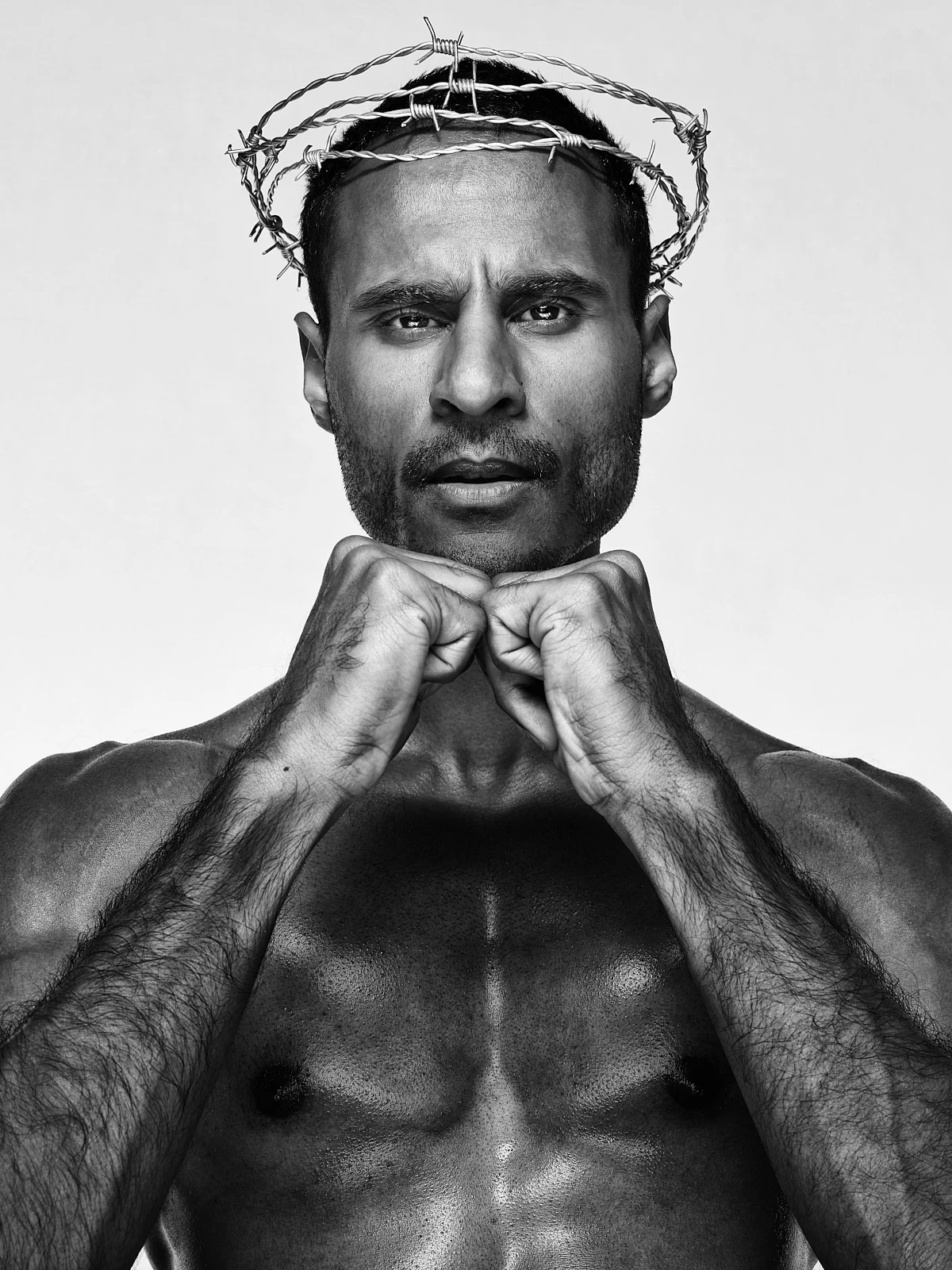 A man with his hands clasped beneath his chin and a woven headpiece.