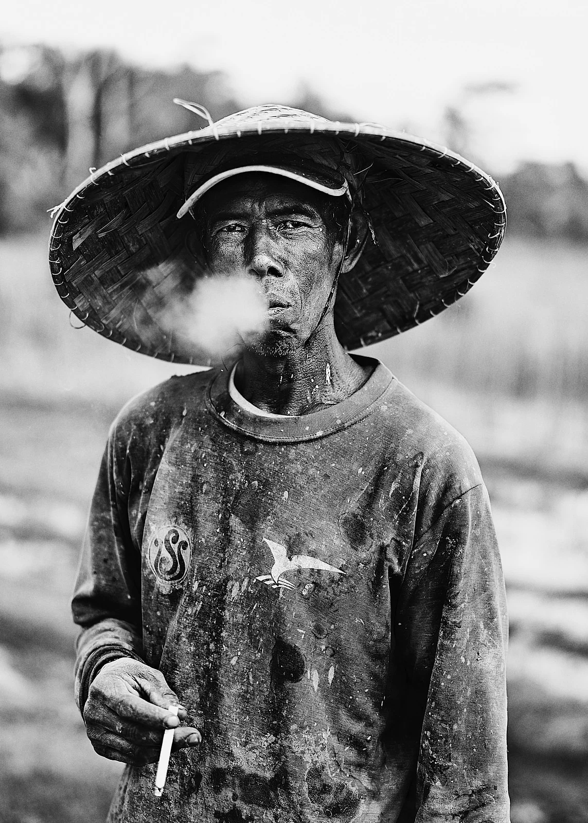 A person in a broad woven hat exhaling smoke outdoors.