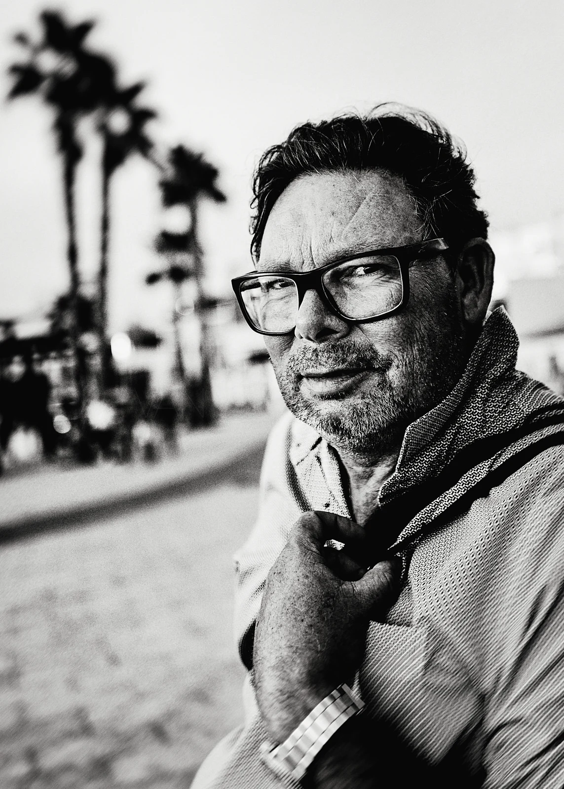 A man with glasses and a scarf standing in a palm-lined street.