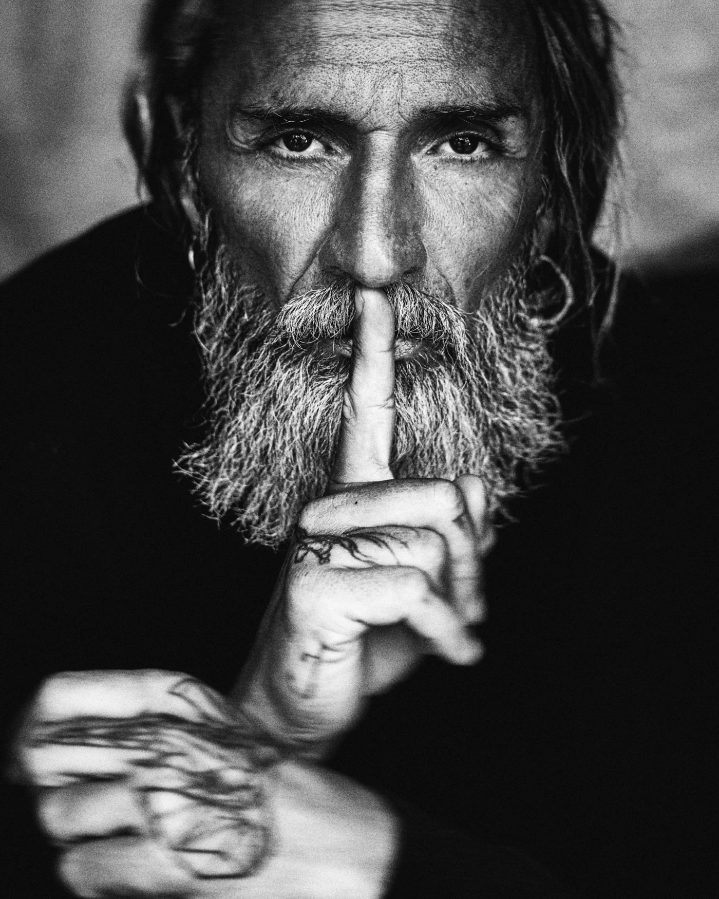 A close portrait of a long-haired, bearded man holding a finger to his lips.