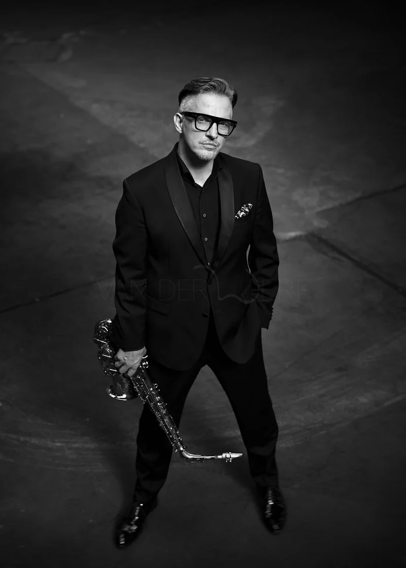 A man in a black tuxedo holding a saxophone, standing on a dark floor, looking confidently at the camera in a black-and-white photo.