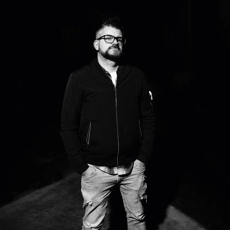 A man with glasses, a beard, and a mohawk hairstyle standing outdoors at night, wearing a black jacket, plaid shirt, and ripped jeans.