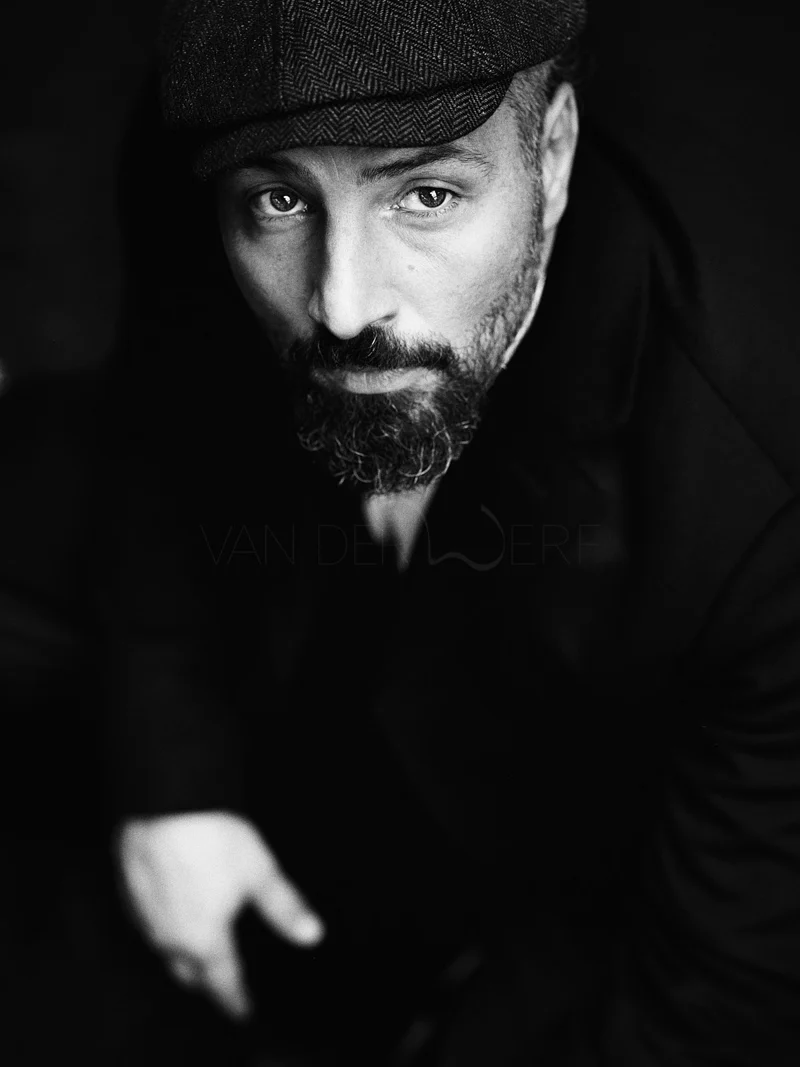 Black and white close-up portrait of a man wearing a flat cap, with a beard and mustache, gazing directly at the camera.